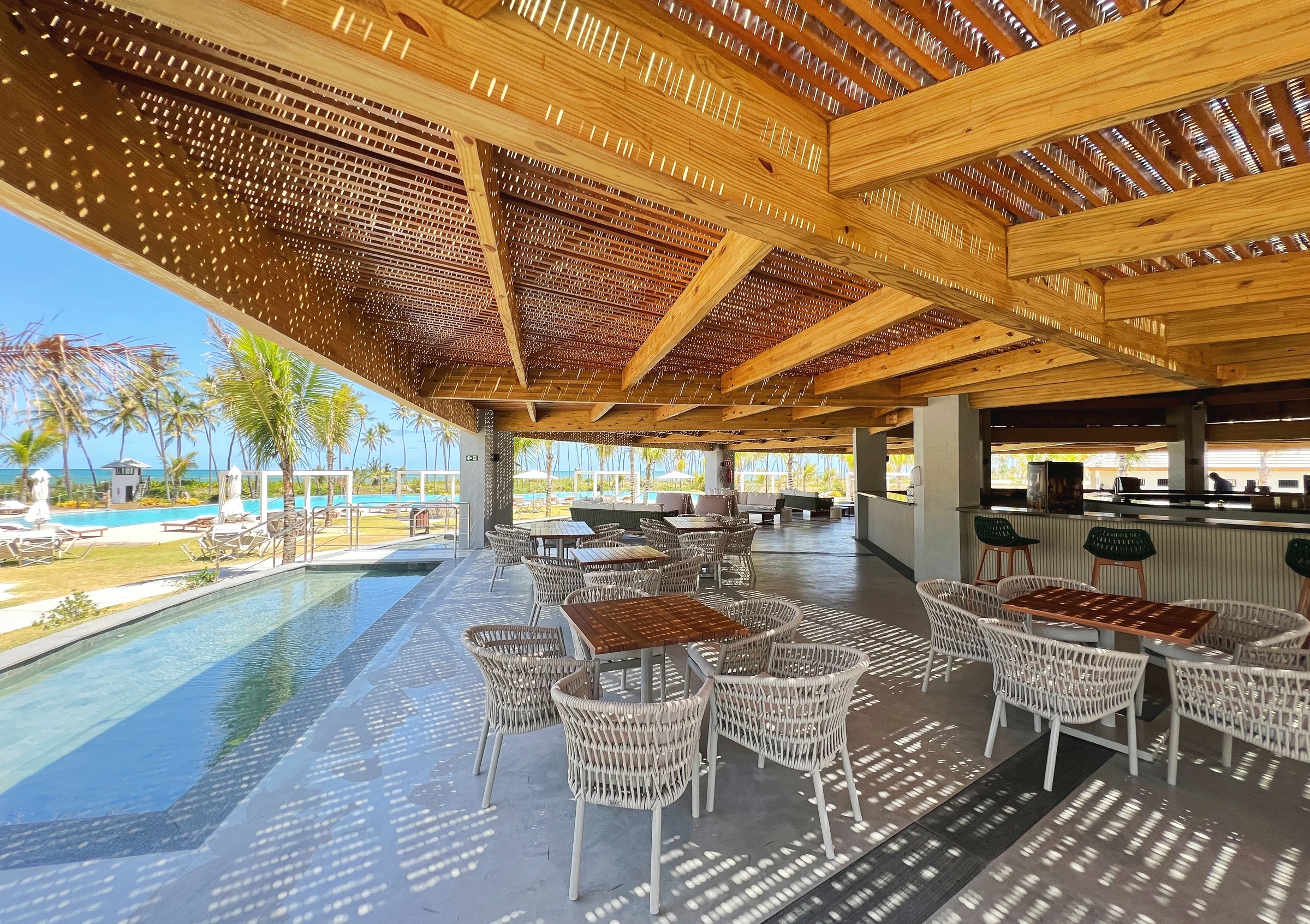 Mesas sob o pergolado do bar, com espelho d’água e piscina ao fundo.
